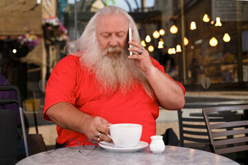 A man with a long, flowing beard is talking on his smartphone while enjoying a cup of coffee in a bustling cafe