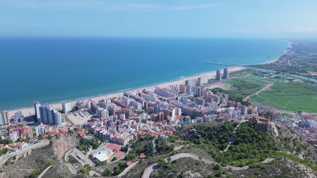 Aerial panoramic view of Cullera, Spain with coastal skyline, Mediterranean Sea, urban buildings, historic castle, and surrounding green farmland under clear blue sky