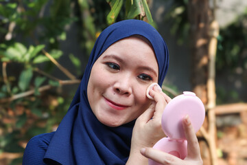 Young Muslim woman wearing hijab applying makeup with compact powder while looking in mirror...