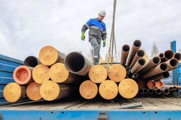 A construction worker in a helmet and gloves inspects steel pipes stacked on a truck. The setting is industrial, showcasing heavy machinery and materials.