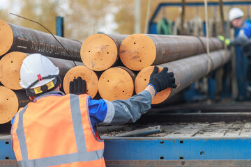 Construction workers in safety gear loading steel pipes onto a truck. The scene depicts teamwork and industrial activity in a construction setting.