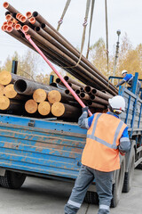 A construction worker in safety gear loads pipes onto a truck at a construction site. The scene captures teamwork and industrial activity.