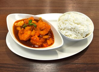 Plate with white rice and sweet and sour tiger prawns in red sauce with vegetables served in white bowls.