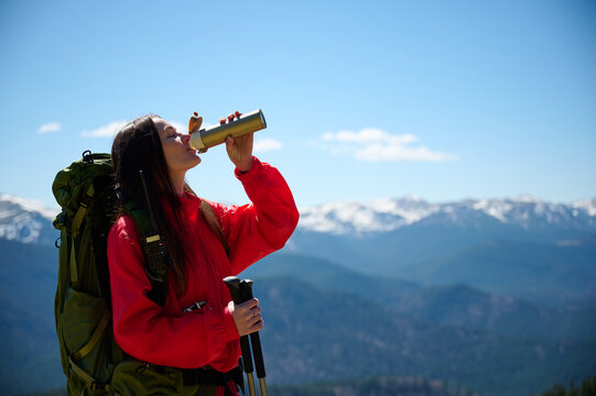 Woman in hiking gear taking a sip of water during mountain hike. Sporty female standing in front of snowy peaks. - Powered by Adobe