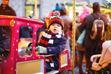 Funny little kid boy on a carousel at Christmas funfair or market, outdoors. Happy child having fun. Traditional xmas market in Germany, Europe. Holiday, children, lifestyle concept..