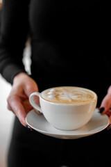 Person holding a white ceramic cup of cappuccino with foam and subtle latte art. Cozy coffee moment in soft light, perfect for lifestyle, cafe, or beverage themes