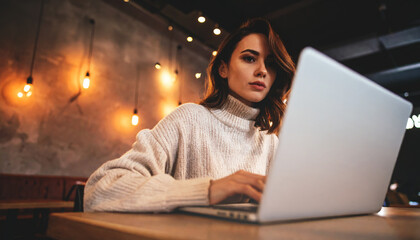 A woman in a cozy knit sweater works on her laptop in a dimly lit stylish cafe with warm pendant lighting.