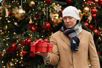Adult caucasian man holding red gift near Christmas tree. Xmas holiday spirit. Cold weather.