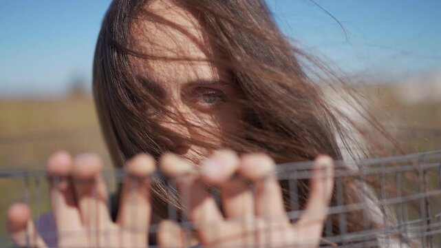 immigration woman reaches forward with her hands placed on a wire fence. The wire stands out in front of the woman, as strands of her hair are caught in the wind.