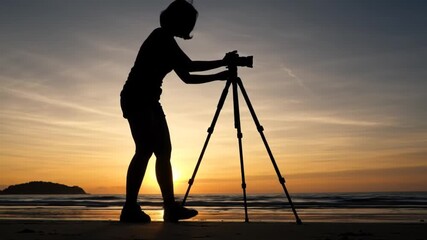 Silhouette of a photographer setting up a camera on a tripod at sunset on the beach with waves crashing - Powered by Adobe