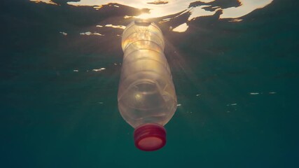 A single-use plastic bottle floating below water surface on sunrays in the sunset time, slow motion, backlit (contre-jour) - Powered by Adobe