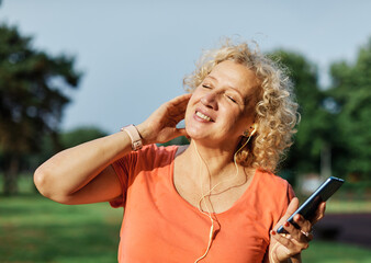 Happy active mid aged woman having fun using smartphone and wearing earphones and sportswear, checking music or an exercise app, after having an exercise sport activity outdoors