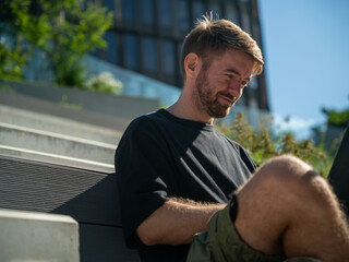 Smiling caucasian man modern urban freelancer typing on notebook remotely in outdoor settings, sitting cross legged on sun-drenched wooden public steps on a summer day.