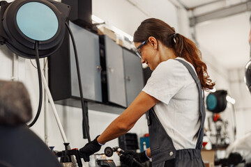 A determined woman wearing overalls is actively using various tools in a modern automotive workshop...
