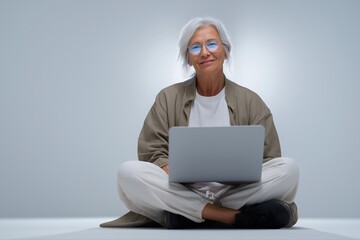 Elderly caucasian woman with laptop in casual attire sitting cross-legged on floor