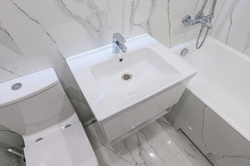 Clean, modern bathroom featuring white fixtures: a toilet, a sink with a chrome faucet, and a bathtub. Marble-patterned tiles on the floor and walls add a touch of elegance