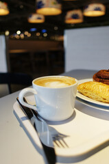 A white cup of cappuccino with latte art sits on a tray with a pastry and cutlery. The scene is in a modern cafe with warm, blurred lights in the background.