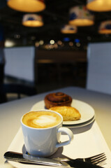 A white cup of cappuccino with latte art sits on a tray with a pastry and cutlery. The scene is in a modern cafe with warm, blurred lights in the background.