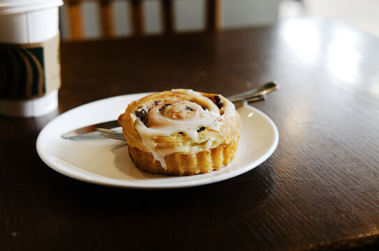  A close-up of a cinnamon roll with icing on a white plate, placed next to a takeaway coffee cup on a dark wooden cafe table.