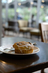  A cinnamon raisin roll with white icing served on a plate with a fork and knife, sitting on a dark wooden table inside a cafe with a softly blurred background.