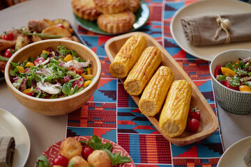 Closeup showing traditional Kwanzaa table setting with grilled corn on cob, fresh salad with greens and vegetables, assorted baked goods, colorful patterned table runner