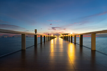 Long walkway floor on wooden structure extending into calm sea at twilight with warm glow of...