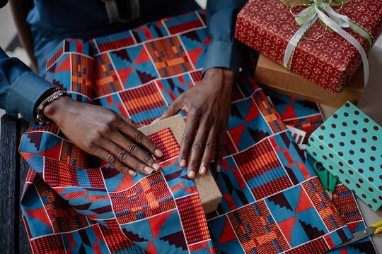 Black adult man wrapping gift box with vibrant patterned fabric, celebrating Kwanzaa tradition, hands arranging present on table surrounded by other wrapped gifts and festive items
