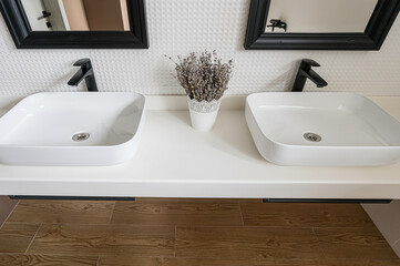 Fototapeta premium A modern bathroom with two square, white sinks, black faucets, black-framed mirrors, a white countertop, and a pot of dried flowers