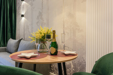 Cozy setup featuring a bottle of champagne, flutes, and yellow flowers on a round table, set against a stylish backdrop with forest-themed wallpaper