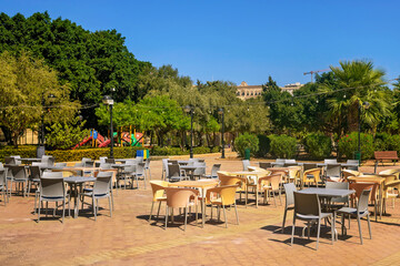 Urban square with yellow and gray tables and chairs in a park with tropical greenery, street lamps and a playground in the background on a sunny day.