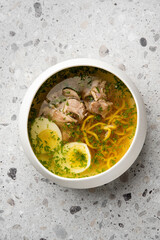 Homemade clear chicken soup with noodles, potatoes, and herbs in a white bowl on a stone surface. Comfort food, top view, rustic presentation, natural lighting