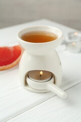 Aroma lamp with burning candle and slice of grapefruit on white wooden table against grey background, closeup