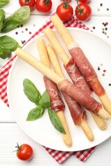 Delicious grissini sticks with prosciutto, peppercorns, tomatoes and basil on white wooden table, flat lay