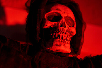 Close-up skull in a hooded cloak under deep red light at night. Spooky Halloween decoration with dramatic shadows and texture.