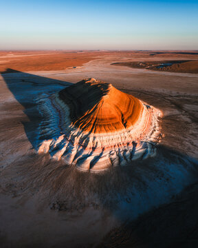 Aerial view of the stunning rock formation, its layers painted in hues of orange and white by the setting sun, casts a long shadow across the arid landscape, Mount Bokty, Mangystau Region, Kazakhstan.