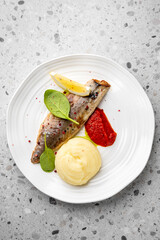 Grilled fish fillet with mashed potatoes, red sauce, spinach leaves, and lemon wedges on a white plate. Gourmet restaurant-style food presentation on textured background