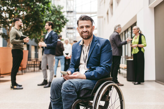 Portrait of smiling male business expert with physical disability sitting on wheelchair while holding smart phone at con