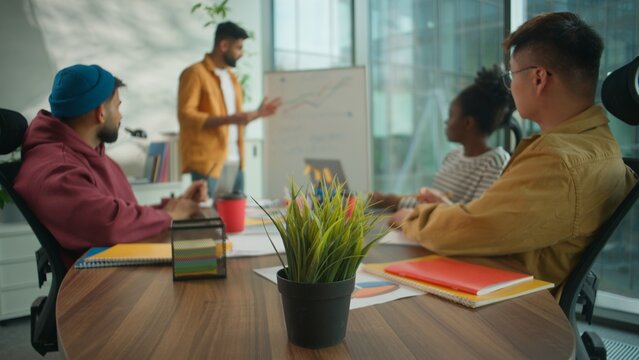 Multicultural informal diversity business team in coworking office listening to male colleague leader presenting financial growth chart on whiteboard discussing startup strategy corporate analytics