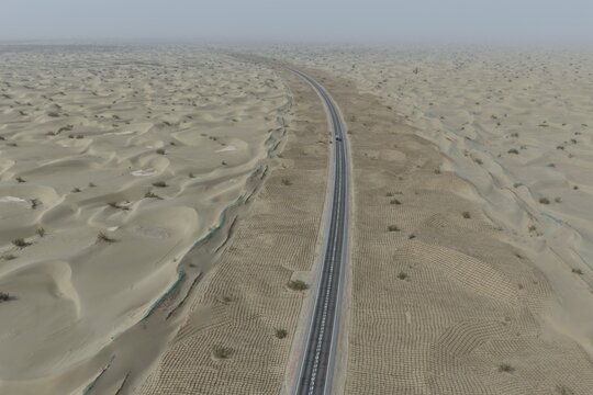 Aerial view of a long road cutting through the vast, undulating ochre dunes under a hazy sky, a testament to human endeavor in the heart of the desert, Taklamakan Desert, China.