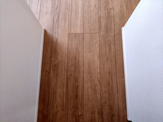 明るい茶色の木目調のフローリングと左右の白い壁が対照的な室内の抽象的な風景です。床中央には点検口のような四角い切れ目が見えます。