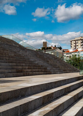 A wide staircase with a glass railing, leading up to a view of a town with a tower and buildings under a blue sky with clouds.