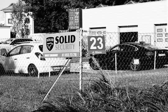 Black and white image of a fenced security lot with parked cars and a visible company sign displaying contact information