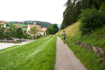 Young man walking along a scenic gravel path in a lush green landscape with charming village in the background