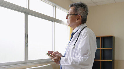 Asian senior male doctor wearing a white coat holding mobile phone and looking outside at hospital. Elderly physician using smartphone to check medical updates.
