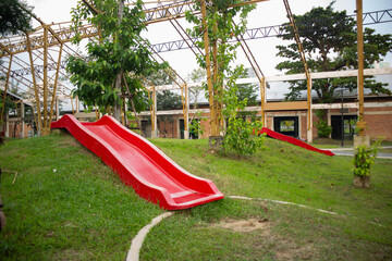 A bright red slide stands on a grassy hill in an outdoor playground beneath a metal frame structure. © BloodysAlice