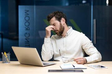 Tired and overworked young businessman sitting at the desk in the office, holding glasses and rubbing his eyes with his hand