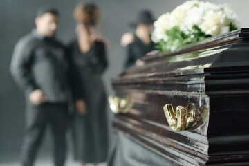 Caucasian man, Black woman, and another adult standing together in background mourning near closed wooden coffin with flowers during funeral ceremony