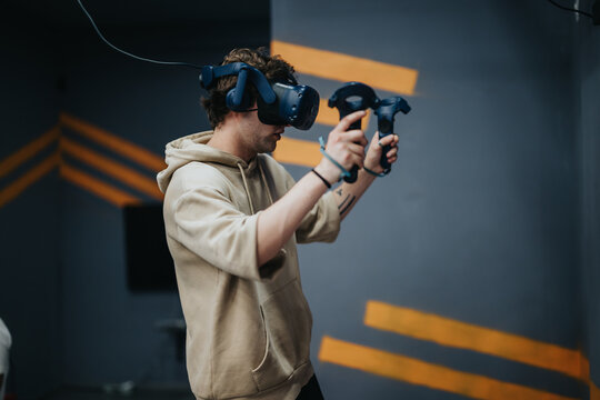 A young man immersed in a virtual reality gaming environment, using head-mounted gear and hand held controllers. Set in a contemporary indoor space, emphasizing interactive entertainment. - Powered by Adobe