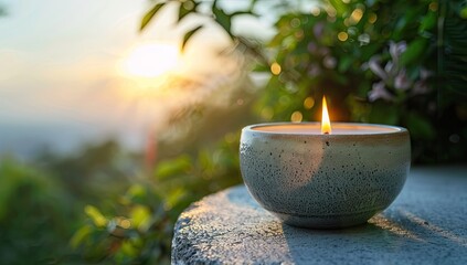 A lit candle in a rustic bowl sits on a stone surface outdoors, bathed in the warm glow of a setting sun with a blurred background of lush greenery