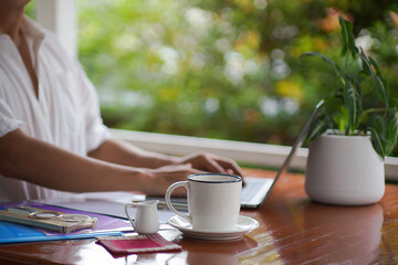 Person working on laptop with coffee cup on wooden desk near window and green plant, cozy home office workspace.